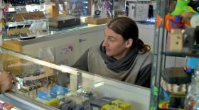 A man wearing a hoodie with a ponytail sits behind a counter. Multiple items are on display in glass cases.