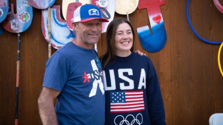 A dad and daughter wearing “Team USA” shirts stand and smile together, with J-shaped signs in the background.
