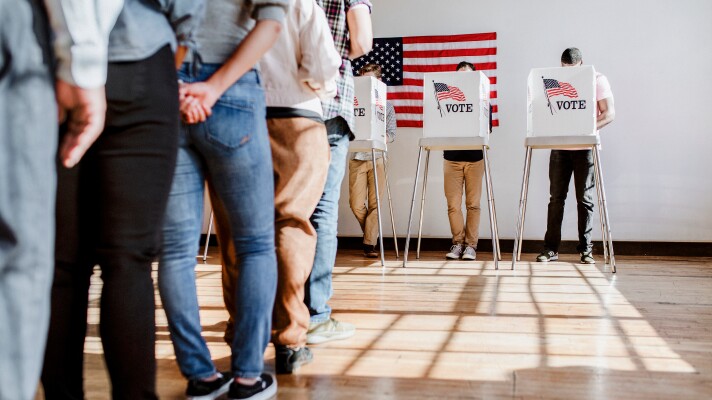 People form a line in front of three voting booths. Each booth is occupied voting by one person. An American flag hangs on a wall behind the voting booths.