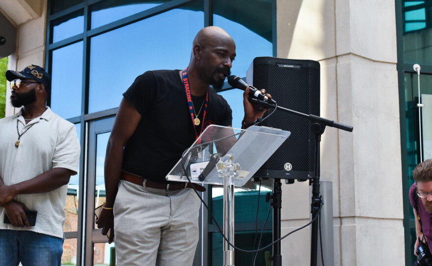 A bald, Black man speaks into a microphone. He wears a black shirt and beige Khaki pants. 