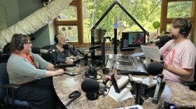 Three people in a studio booth with microphones and computers in front of them.