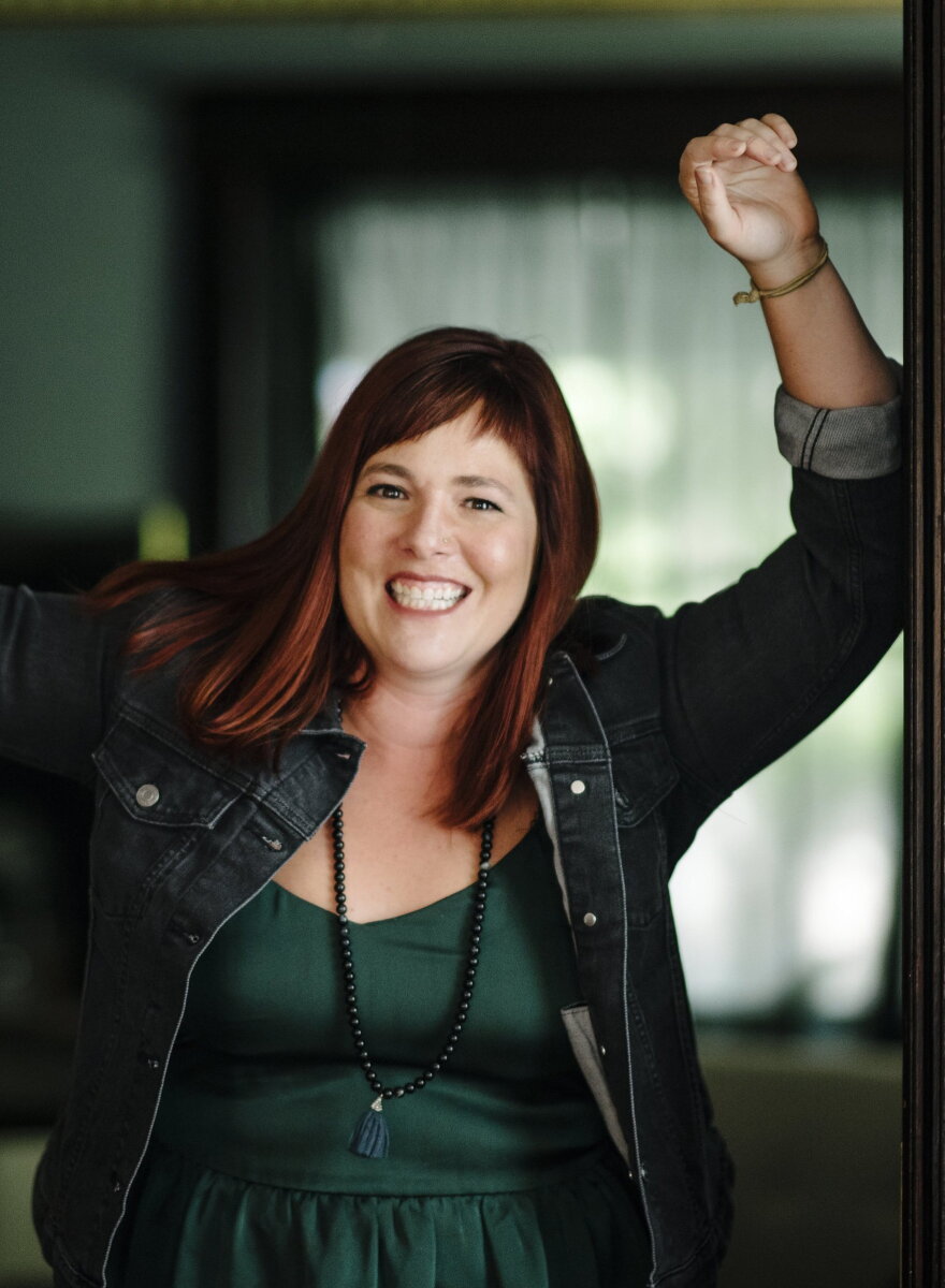 A woman with red-dyed hair leans against a wall with her arm up, smiling.