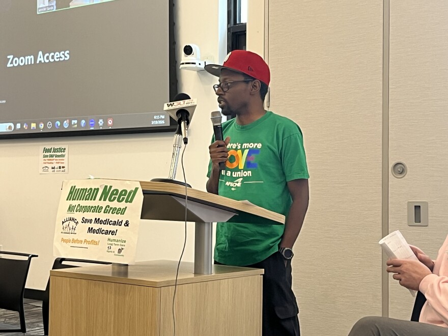 A man with a microphone stands at a podium in the Bloomington Public Library. He wears a ball cap and a pro union shirt