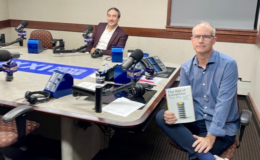 Two smiling men sitting at a table in a radio talk studio: the man back left has short dark hair and a dark mustache and is wearing a white button-down shirt under a burgundy suit; a man front right has very short dark hair and is wearing a blue button-down shirt, jeans, blue socks, and white and red sneakers. He is holding a book titled "The Age of Extraction." 