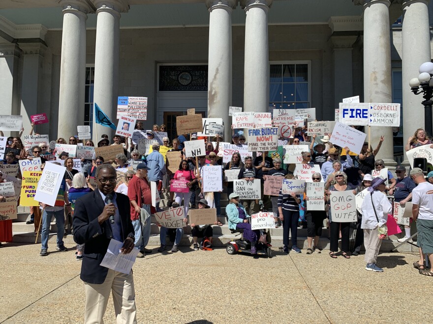 A photo of a man in a jacket holds a microphone as he speaks in front of a crowd of protesters holding signs. Signs read: "FIRE: Cordelli, Osborne, Ammon" and "OMG GOP WTF?" and "Fair Budget Now"