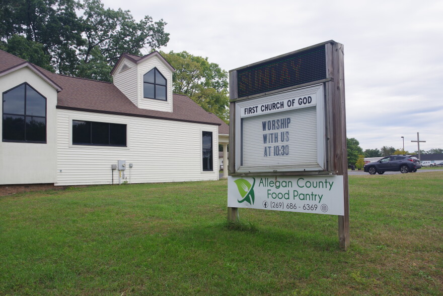 A sign stands in a green grass lawn and in front of a white trimmed wooden building. The wooden post sign has three parts, a digital sign that displays orange text reading "Sunday" at the top, a middle area that has a frame reading "First Church of God" that frames another white sign that reads "Worship with us at 10:30," and finally is a long and thin sign near the bottom that reads "Allegan County Food Pantry." 