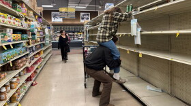 A man hoists a woman on his shoulders to reach water bottles on the top shelf of an otherwise empty set of shelves.