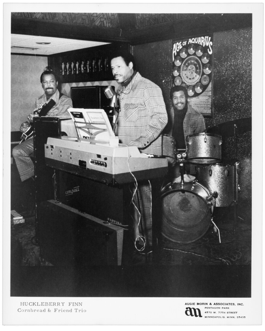 A black and white photo of a trio on stage. On the left a seated man plays guitar, in the middle a man stands behind a organ, and in the back a man sits behind a drum kit. 