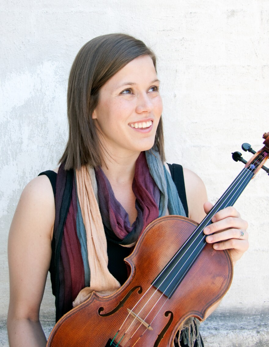 A fair-skinned woman with brown hair and a multi-color loop infinity scarf holds a violen and looks off the the right with a smile. 