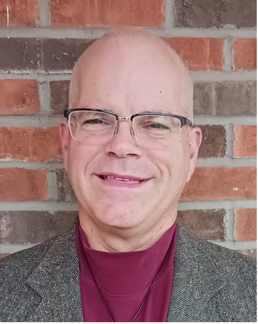 man in maroon shirt with tweed jacket from shoulders up smiles with a brick wall behind