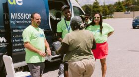 Three volunteers, one woman and two men, dressed in matching green t shirts are talking with a dark-skinned woman in a t-shirt and shorts. They are standing in front of a blue van.