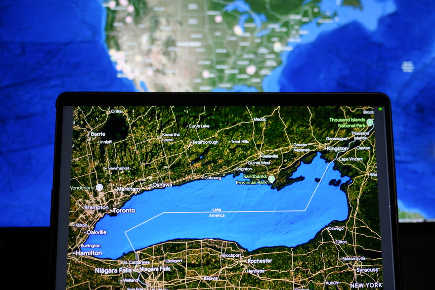 A hand holds up an i-pad that shows a map that is zoomed in on Lake Ontario, which is labeled "Lake America" on the map. Behind it is a computer screen showing a map of the United States. 
