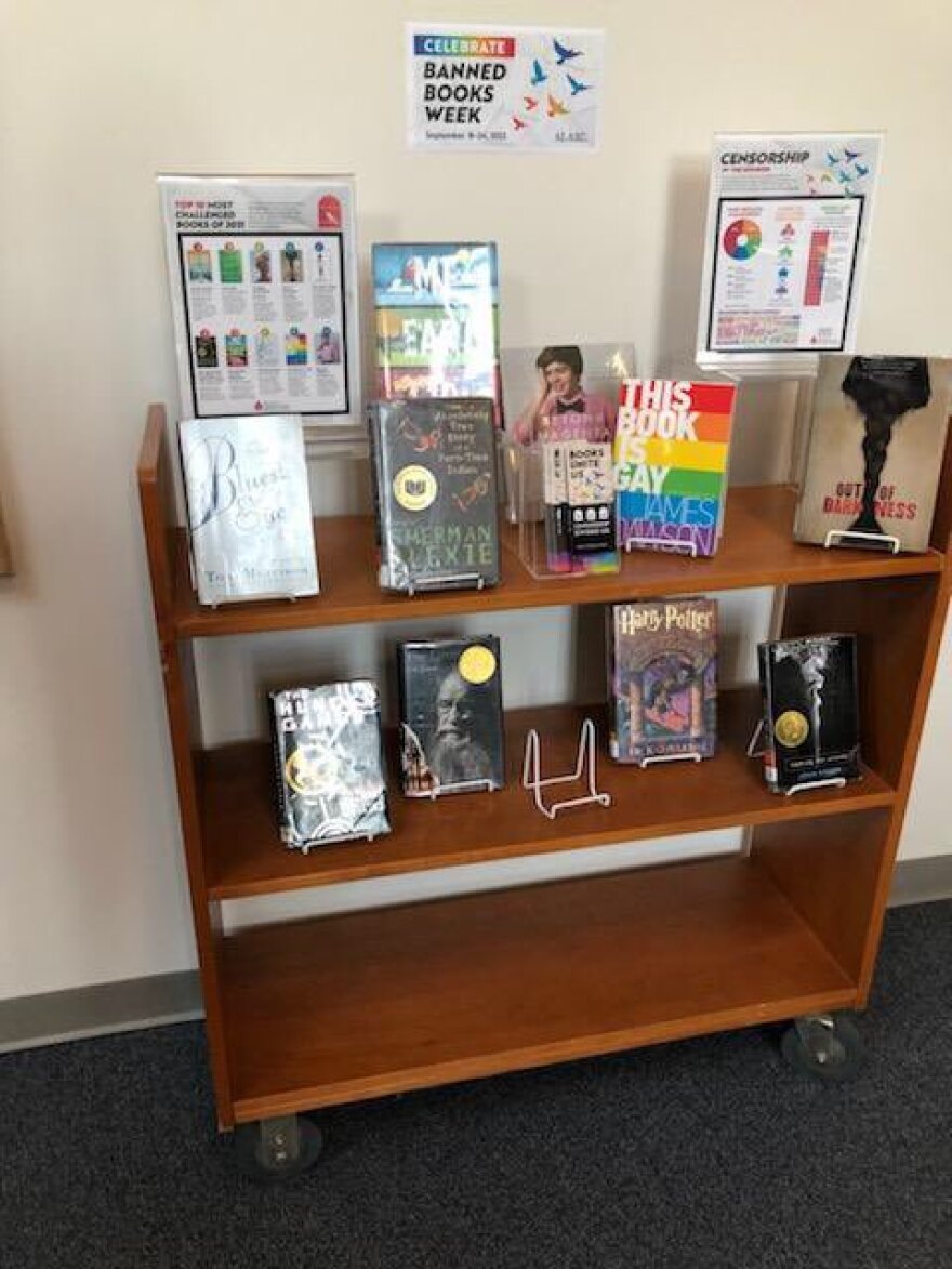 A book shelf under the banner "Banned Books Week" holds copies of "The Giver" by Lois Lowry; "This Book is Gay" by James Dawson; and "The Bluest Eye" by Toni Morrison.