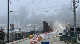 Fencing blocks off the right lane of a bridge while the left side is open. Sailboats float in water on the left side of the image.