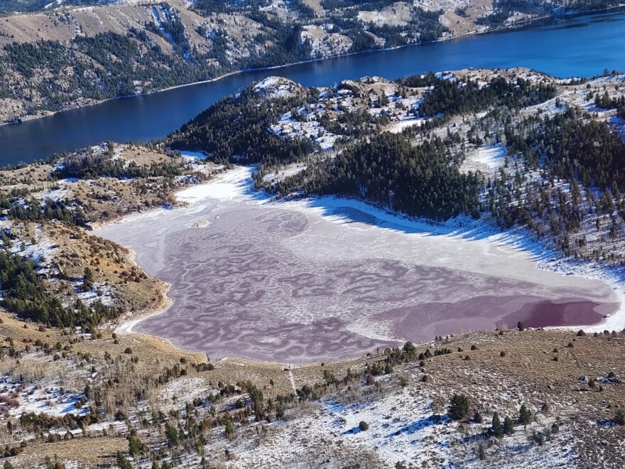 An aerial image of a large blue lake in the background, with a frozen, red lake in the foreground, surrounded by trees, sagebrush and light snow.