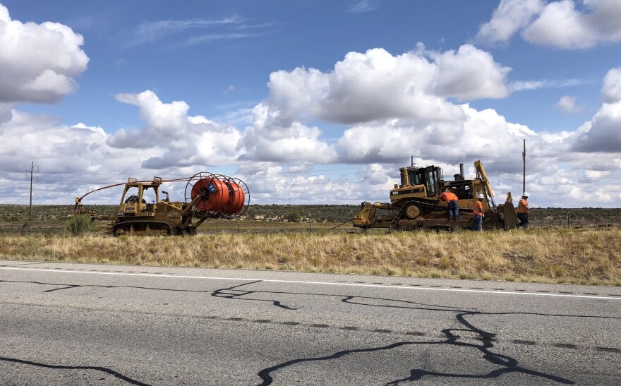 A heavy machine holding a spool of orange cable drives along the highway behind a tractor. 