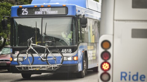The front of a blue bus travels toward the camera on a city street.  On the front, the route marquis reads "25 Troost 83rd and Troost."