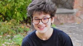 Ellie Lin is sitting on a concrete bench wearing a glasses, a black t-shirt and blue jeans. There is grass and cobblestones behind her. 
