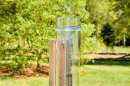A clear plastic rain gauge stands in a summer garden, supported by a wooden post.