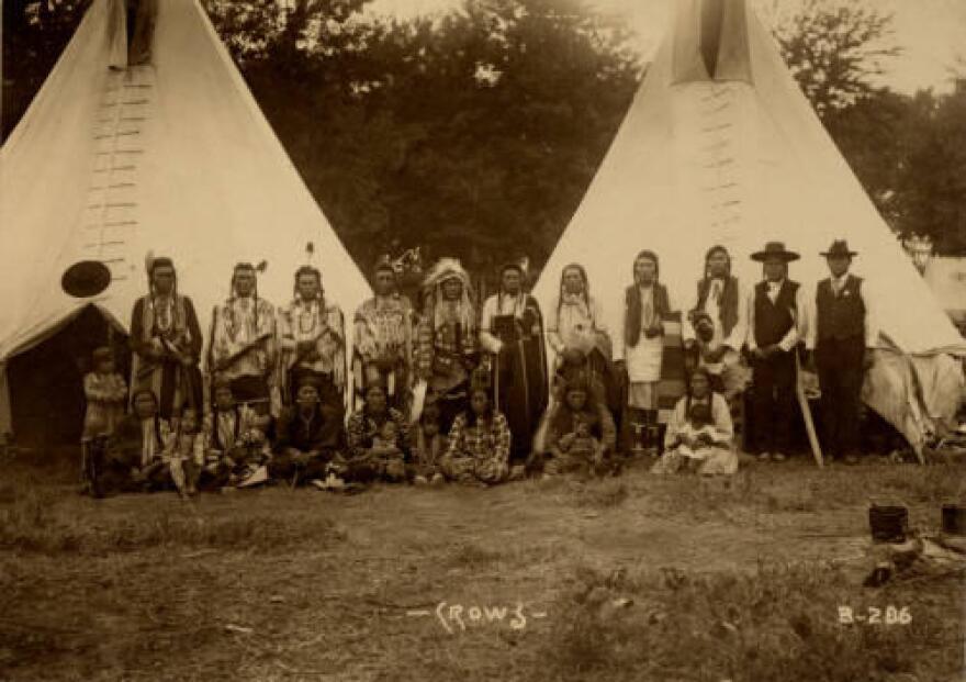 Black-and-white photo showing group of 25 Native American men, women, and children in front of two tipis