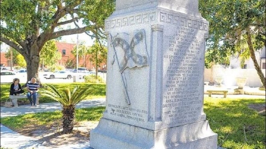 William Loring monument, etched in it is a confederate flag, in a park, with two people sitting in a bench behind it.