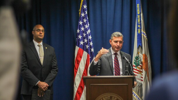 A man in a suit and tie speaks at a podium, with a U.S. flag and another flag behind him. The second flag has a banner that reads "Southern District of Ohio." A second man in a suit and tie stands to the side, hands folded. 