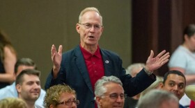 Man standing in a crowd in a meeting room while wearing a dark sports jacket and red collared shirt and extending his hands as he speaks. 