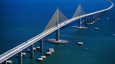A tall bridge with yellow steel supports is surrounded by blue water. A few cars line the bridge, and in the water below are bumpers and rock islands surrounding the bridge. 