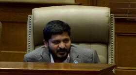 A man with short dark hair and a beard sits in a large, beige leather chair, speaking at a wooden desk in a formal setting.