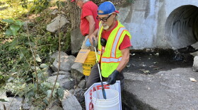 Two middle-aged men in red shirts use trash grabbers to pick up trash in a concrete culvert. 