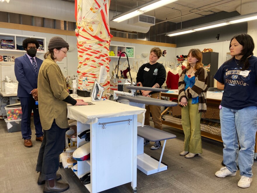 A woman in glasses and a beanie and oversized shirt stands at an iron. Several young people watch as she demonstrates pressing open a seam.