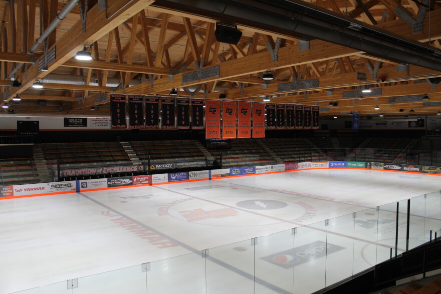A hockey rink with light wood rafters. From the rafters, four large orange banners representing state championships hang with six banners representing state appearances on either side.