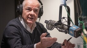 A man inside a radio studio sits behind a microphone talking and gesturing with his right hand.
