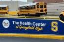 A sign announces the Health Center at Springfield High. Yellow school buses line up behind it.