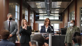 Governor Gretchen Whitmer speaking in front of a crowd at a community center. She speaks at a lectern with two microphone in front of her. There is one woman on her left, our right, wearing a mask facing the crowd. On the other side is Whitmer's American Sign Language interpreter and a man wearing a mask.