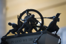 A close up of a mini model of people on top of a platform that reads "In God We Trust." The figures on top of the platform are pulling and pushing a hoop.