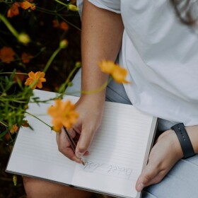 Person sitting on ground near flowering plant and writing 'bucket list' in a lined notebook in lap