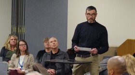 A man in a long sleeve black turtle neck and dark beige pants faces towards the camera as he speaks at a small podium, holding a prepared statement on a sheet of paper. He's facing a row of people, the KRESA board of education. Attendees sit behind him, looking at him as he speaks to to the board