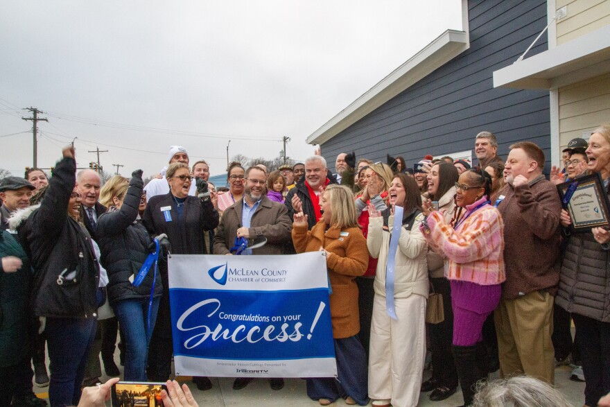 A large group of people cheers as a blue ribbon is cut with a giant pair of scissors.