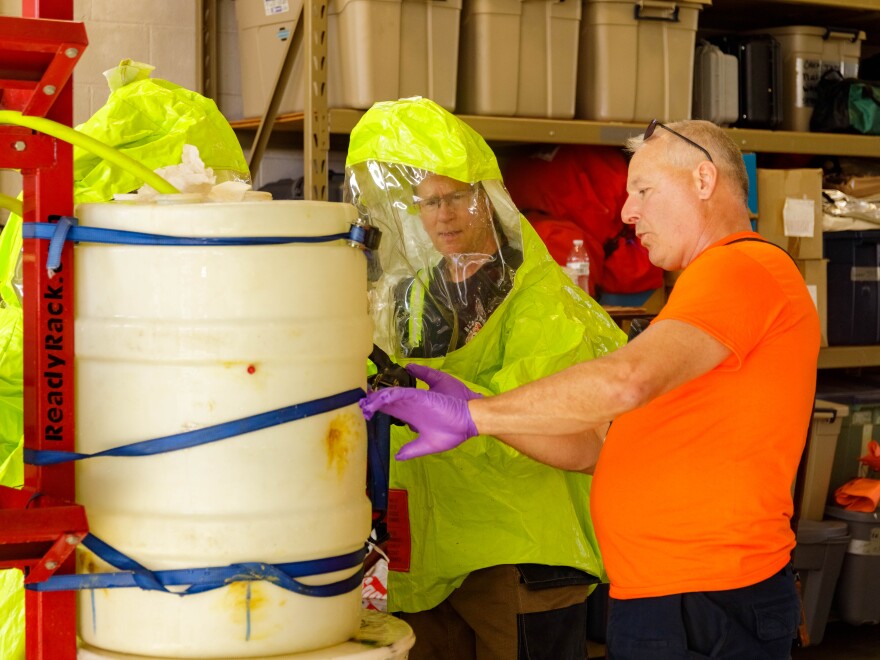 Fire Ops 101 participant is wearing a yellow hazmat suit and going through the hazardous waste cleanup simulation with the help of a Bloomington Local 49 firefighter wearing an orange shirt and purple protective gloves. The simulation includes a leaking tub strapped to a pole.