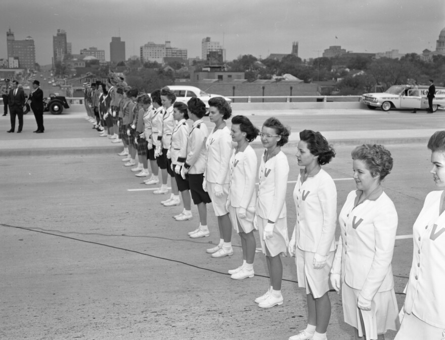  High school girls in uniform form a line across the highway facing south. A few men in dark suits are milling about. Beyond, a few buildings making up the 1962 Austin skyline. 