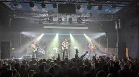 Five band members play on a brightly lit stage in a crowded concert venue.