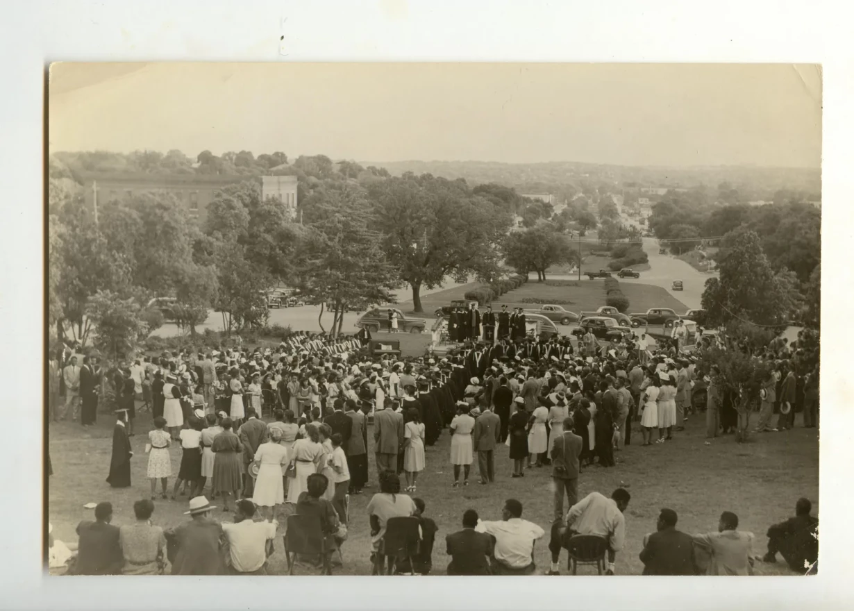 This black and white photograph captures a large gathering of people attending a convocation at Samuel Huston College in 1947. The event is outdoors, set on a grassy hillside with a clear view of rolling hills and a few buildings in the background. The audience is focused on a group of speakers and dignitaries standing around a lectern, with rows of seated uniformed band members in the foreground. Several vintage cars are parked in the vicinity.