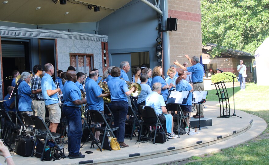 The band director raises a baton to lead musicians playing brass and woodwind instruments on the outdoor stage.