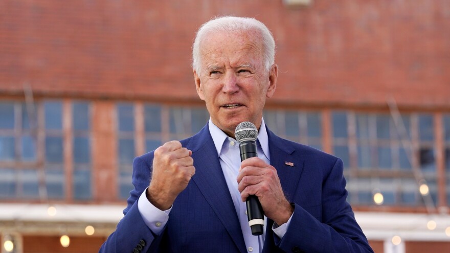 Joe Biden speaking with a micrphone in one hand, and a fist raised. Squinting his eyes while speaking. A large window in the background.