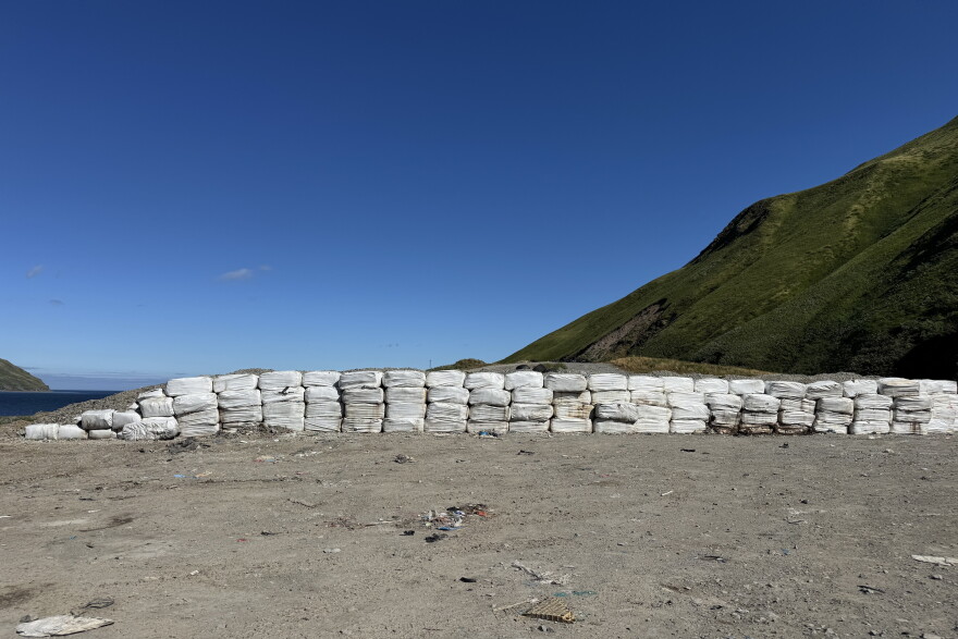 off white cubes are stacked on flat ground covered in dirt. green mountains are in the background