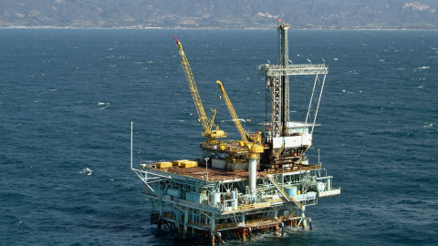 A large offshore oil platform is seen off the California coast. Populated areas and mountains are seen in the distance.