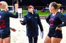 a beach volleyball coach in a black nike jacket and long pants carrying a clipboard talks to two players in black tops with red numbers and b lack shorts on a sandy beach volleyball court