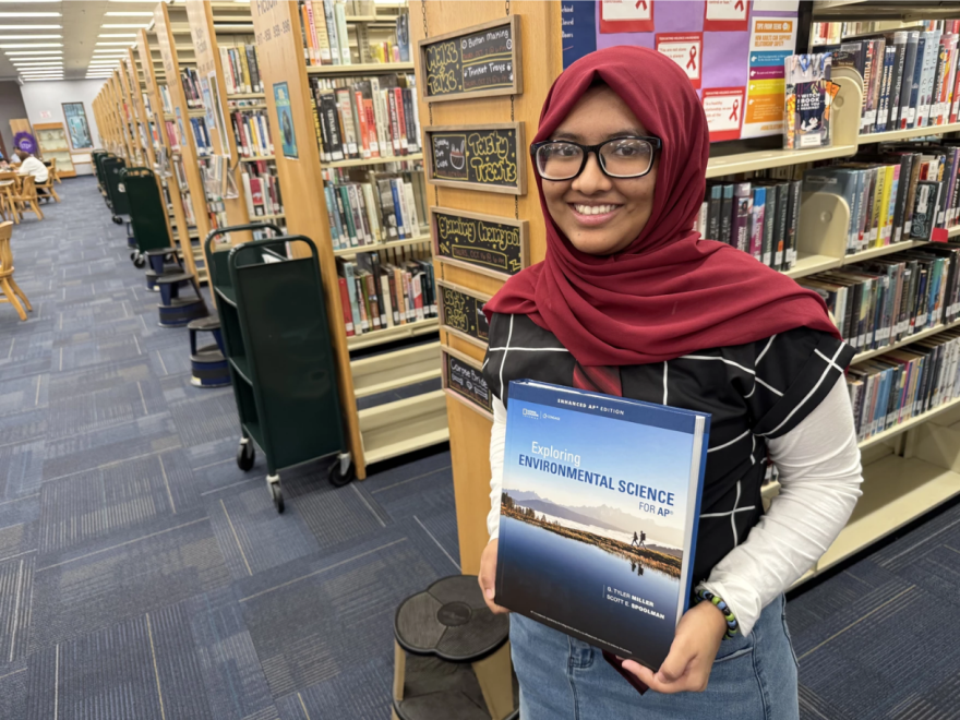 Mujer joven con hiyab rojo y gafas negras sonríe mientras sostiene un libro de texto frente a ella, con filas y filas de estanterías llenas de libros a sus espaldas.
