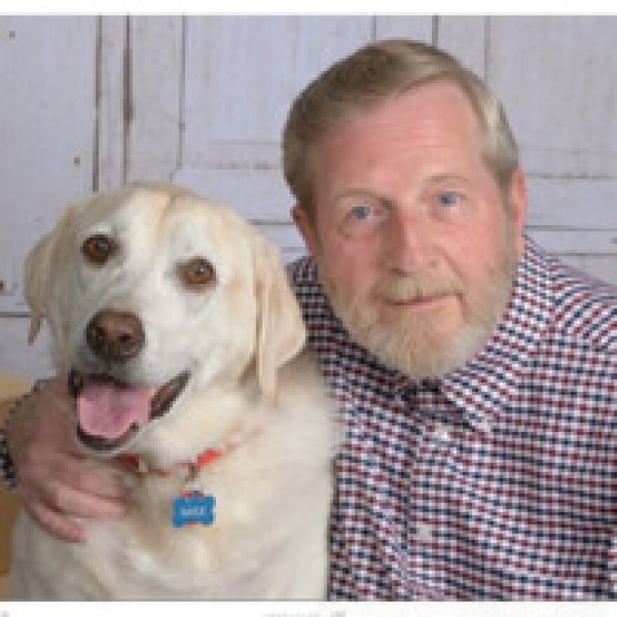 A man with gray hair and a full beard, wearing a red and blue plaid button-down shirt, smiles for a portrait alongside a yellow Labrador retriever with its tongue out, both photographed against a white paneled background.